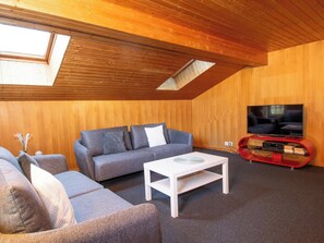 Ein gemütliches Wohnzimmer mit schrägen Wänden, einer Sofagarnitur, einem Couchtisch und einem Fernseher an einer Holzwand.