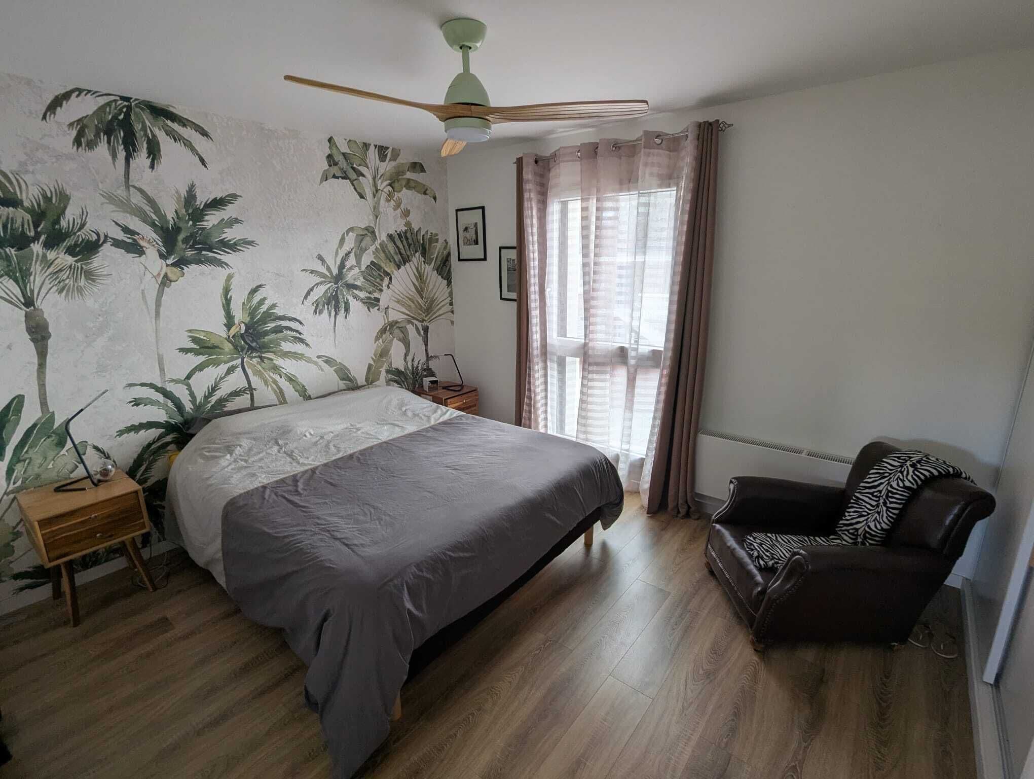 Une chambre à coucher avec un lit, une chaise, un ventilateur de plafond et une fenêtre avec des rideaux.
