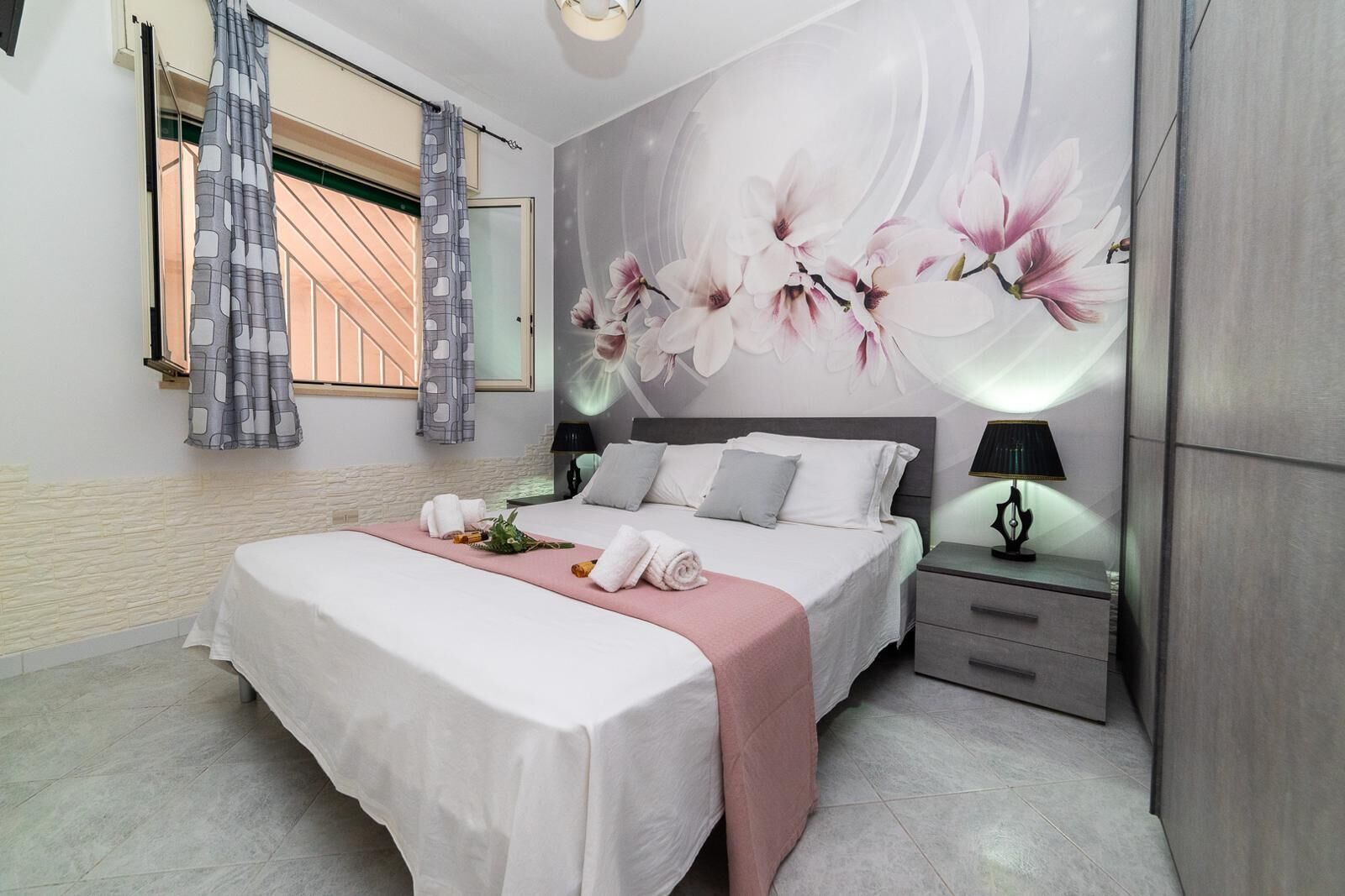 A bedroom with a bed, a nightstand, a lamp, a window with curtains, and a wall mural.