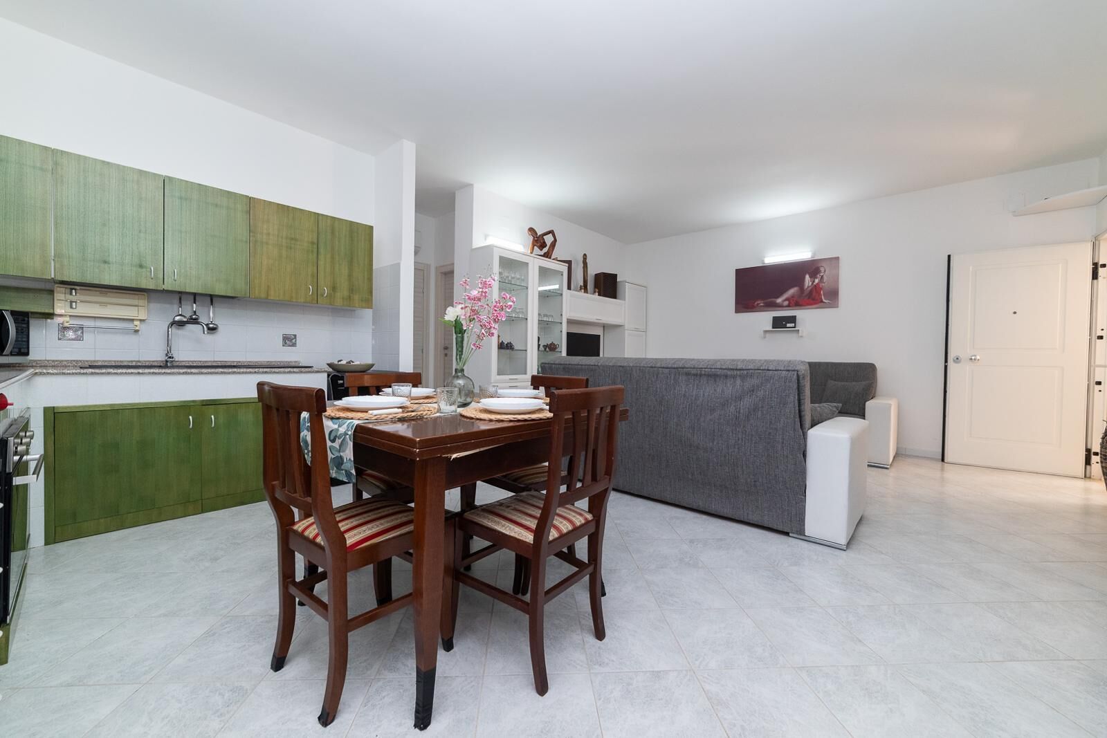 Une cuisine moderne avec des meubles de couleur verte, une table à manger dressée pour quatre personnes et un canapé gris dans l’espace salon.