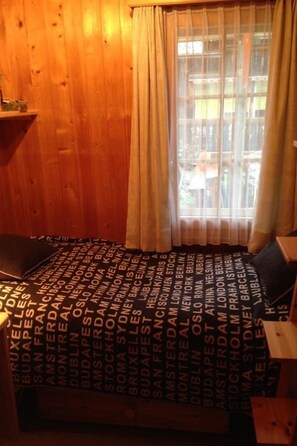 Quarto com parede de madeira e janela com cortinas. Há uma cama com colcha estampada.