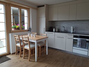 A kitchen with wooden cabinets, a dining table with chairs, a window with flowers, and a built-in oven. - Sonnen-Panorama Appartement/Fewo, Dusche, WC 1-Zimmerwohnung by Interhome (Adelboden)
