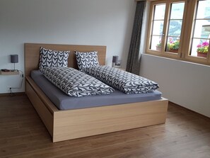 A modern bed with a wooden frame and a grey mattress, two heart-patterned pillows, a bedside table with a lamp, and a window with a view of greenery. - Sonnen-Panorama Appartement/Fewo, Dusche, WC 1-Zimmerwohnung by Interhome (Adelboden)