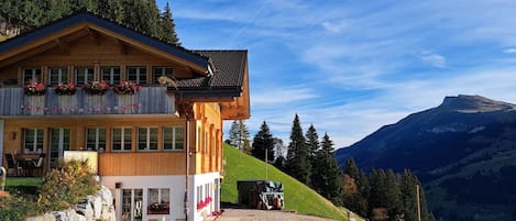 Un chalet de madera con balcón, camino de grava y montañas al fondo.