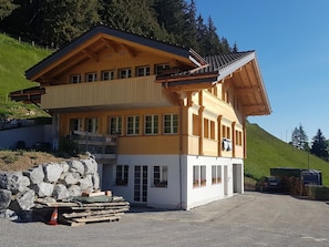 A two-story building with a sloped roof, multiple windows, and a stone wall in front. - Sonnen-Panorama Appartement/Fewo, Dusche, WC 1-Zimmerwohnung by Interhome (Adelboden)