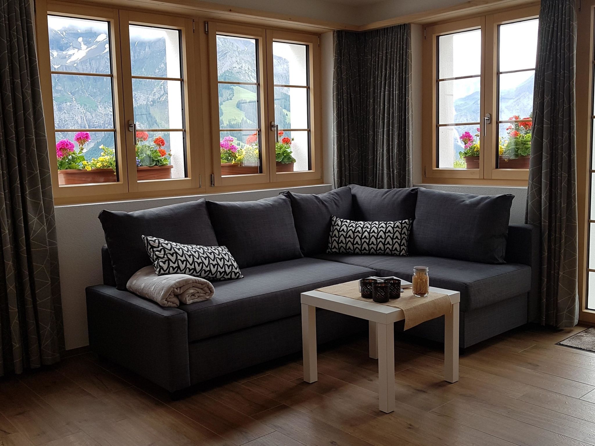 Ein modernes Wohnzimmer mit einem dunkelgrauen Sofa, zwei gemusterten Kissen, einem kleinen weißen Tisch und Blick auf Berge durch große Fenster.