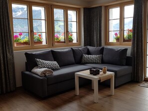 A modern living room with a dark grey sofa, two patterned pillows, a small white table, and a view of mountains through large windows. - Sonnen-Panorama Appartement/Fewo, Dusche, WC 1-Zimmerwohnung by Interhome (Adelboden)