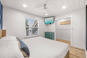 Une chambre à coucher comprenant un lit, un téléviseur, une fenêtre avec des stores, une commode et une porte.