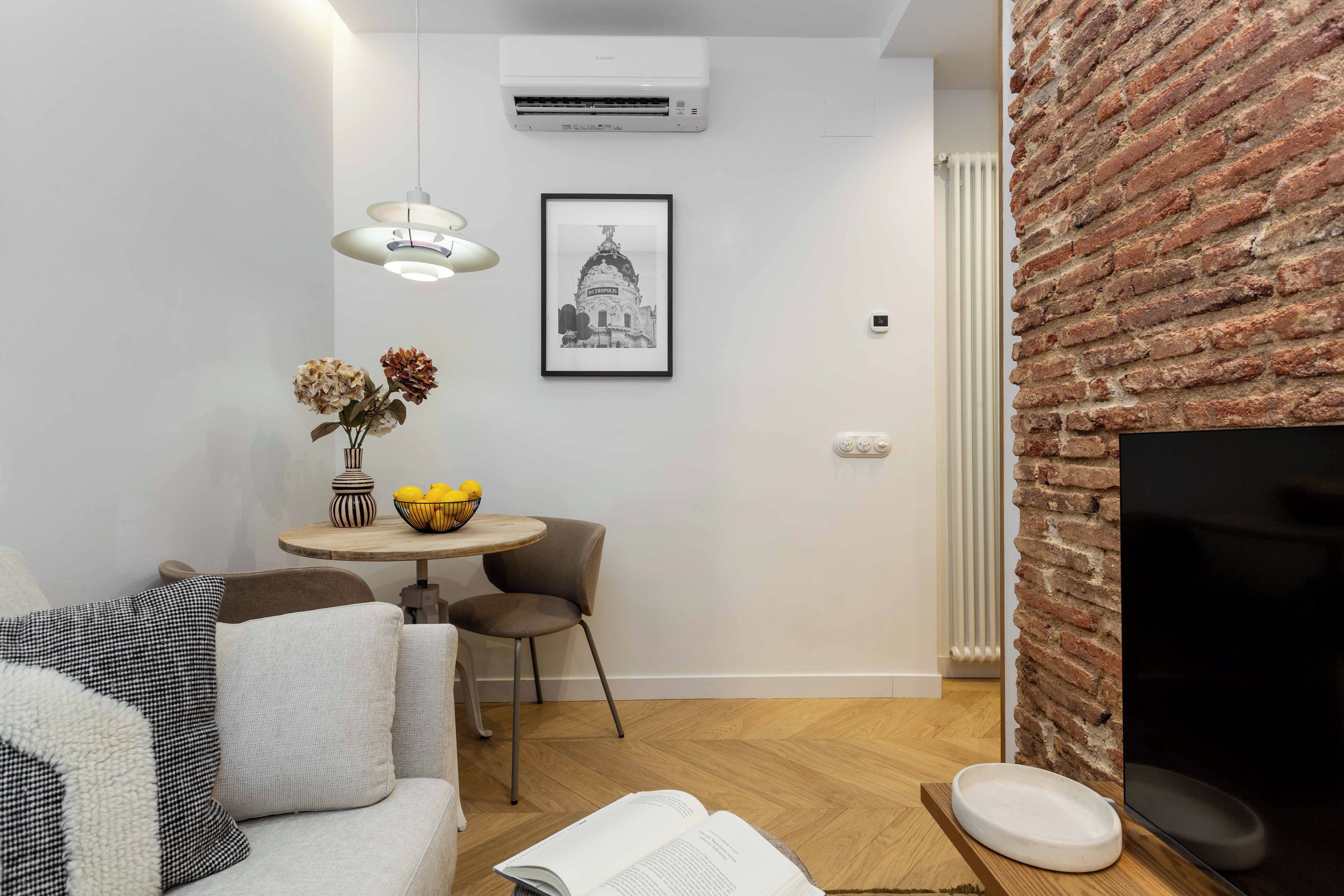 Un espacio habitable compacto con un sofá, una mesa de comedor con un tazón de fruta, un cuadro en la pared y una pared de ladrillo decorativa.