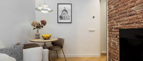 Un espacio habitable compacto con un sofá, una mesa de comedor con un tazón de fruta, un cuadro en la pared y una pared de ladrillo decorativa.