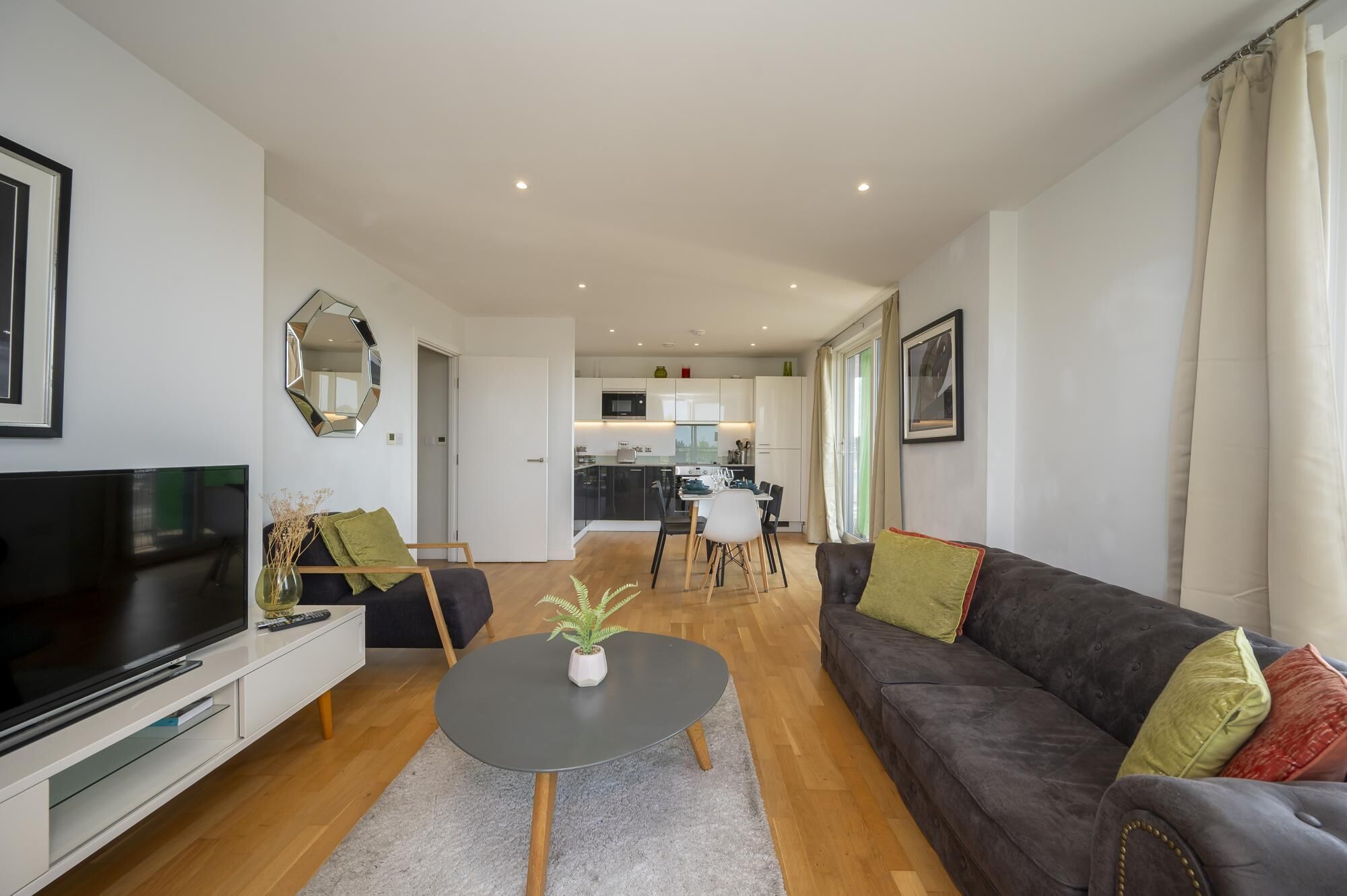A modern living room with a flat-screen TV, a sofa, a coffee table, and a dining area in the background.