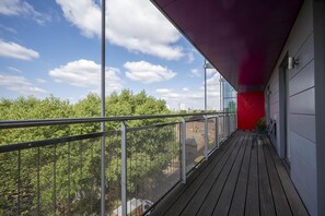Ein Balkon mit Metallgeländer, einer roten Wand und Blick auf Bäume und Gebäude.