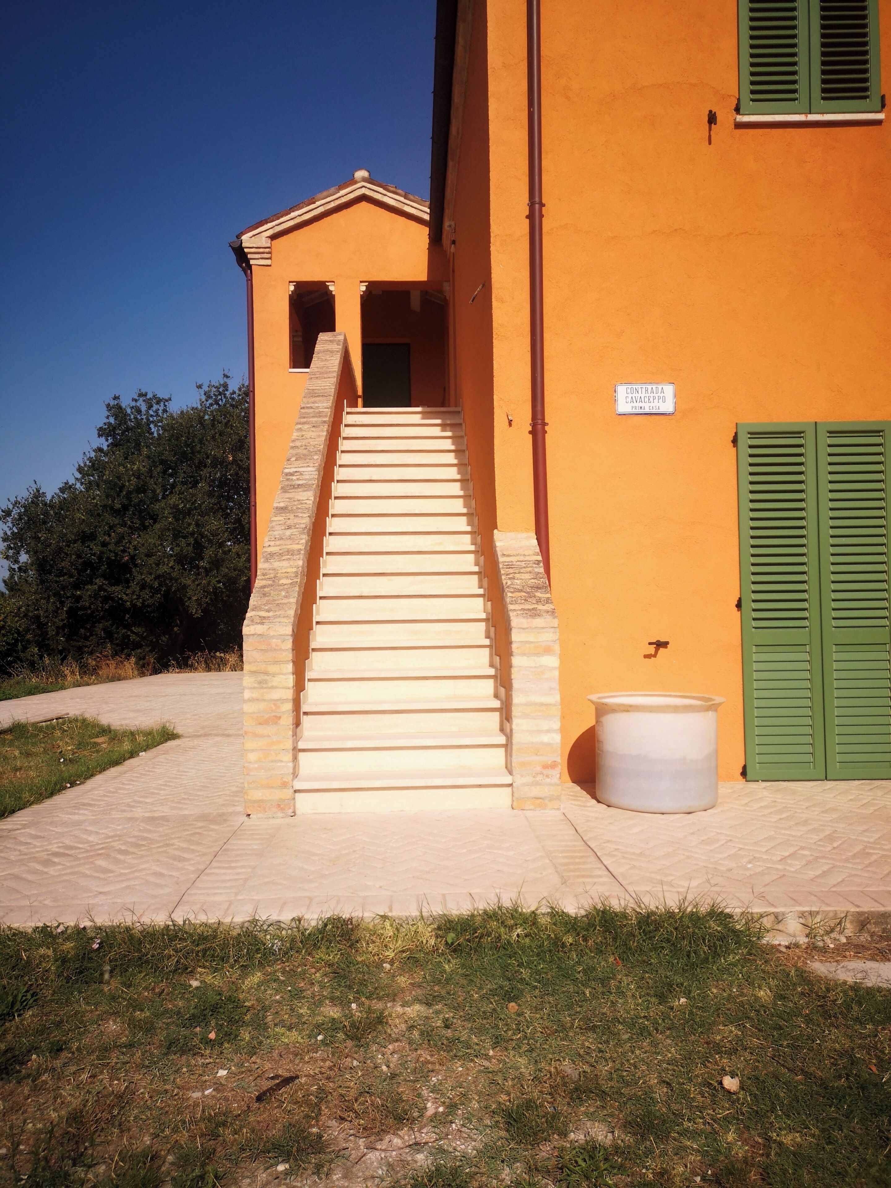 Un edificio con una scala che conduce a una porta, una finestra con persiana verde e un cartello appeso al muro.