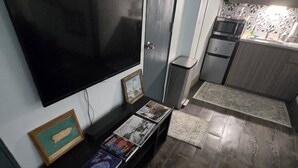 A living room with a flat-screen TV mounted on the wall, a small shelf with books and a framed picture, and a kitchen area with a microwave and a sink. - Beach House Vega Alta PR (Sabana)