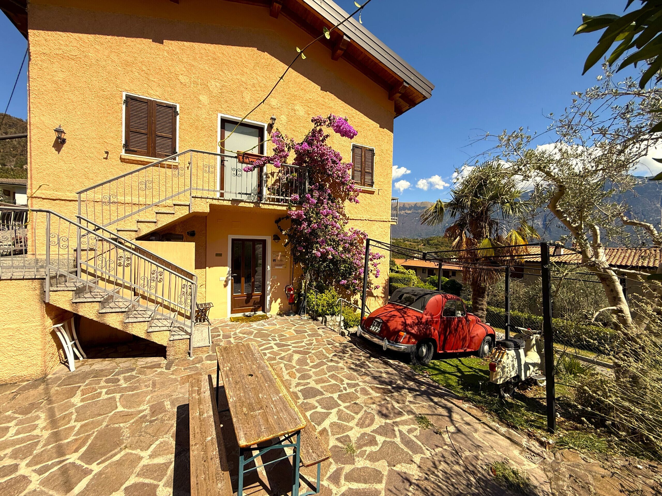 A yellow building with a stone staircase, a wooden bench, and a red car parked outside.