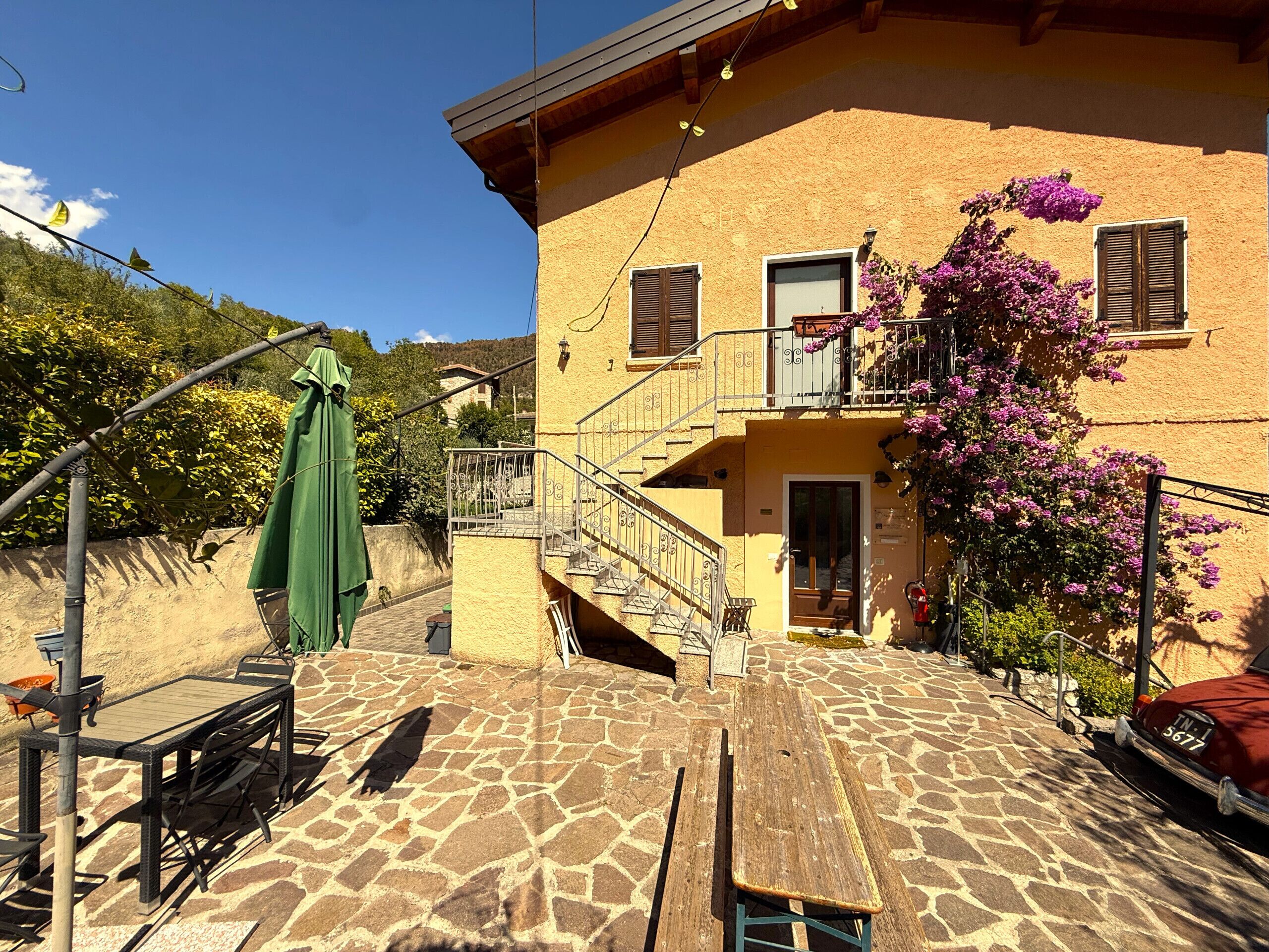 A building with a stone patio, outdoor seating, and a staircase leading to a balcony with purple flowers.