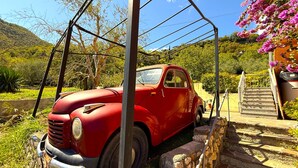 Une voiture rouge d’époque est garée dans un espace délimité par des murs de pierre, avec un escalier menant à un bâtiment orné de fleurs roses.