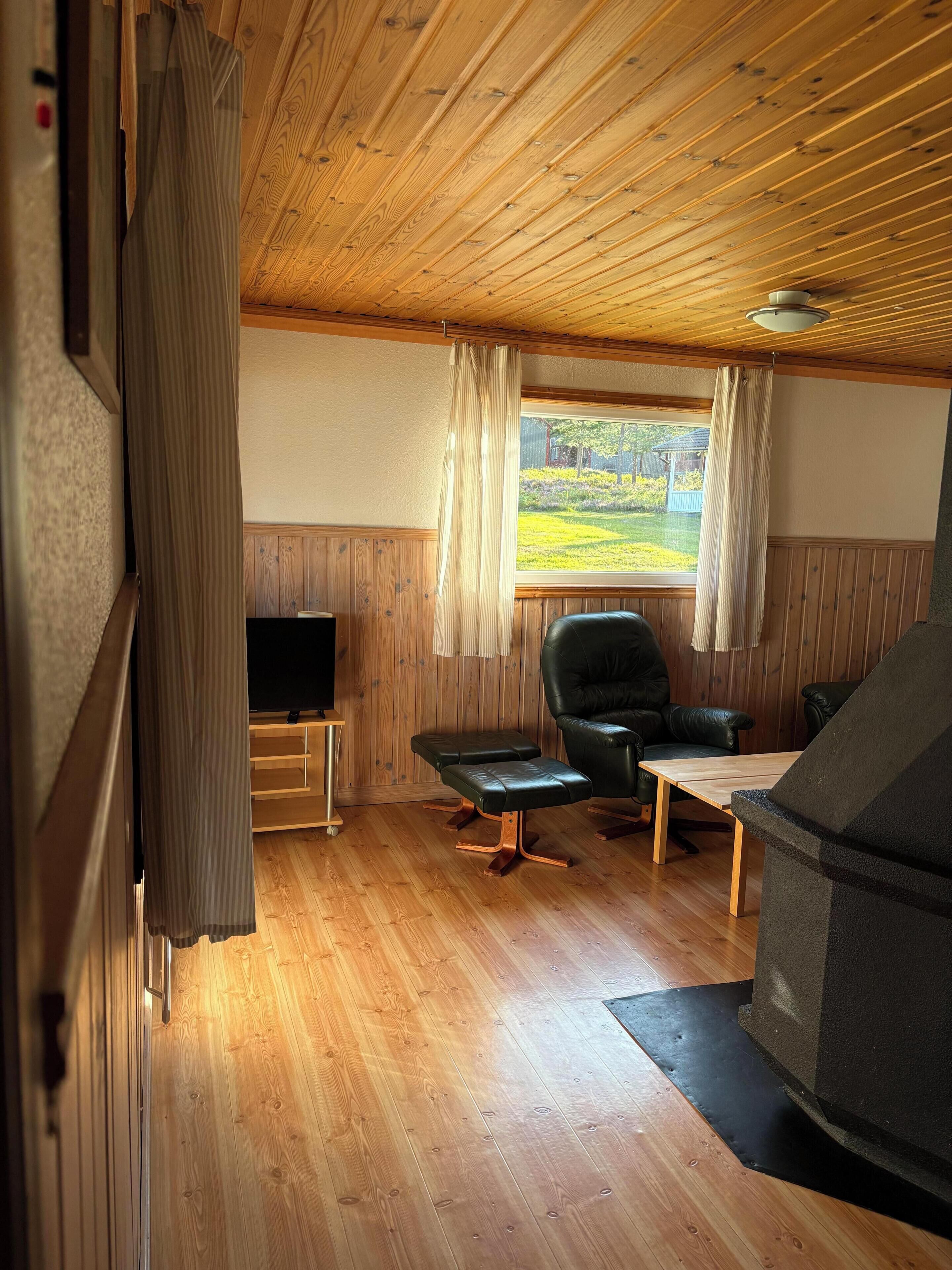 A cozy room with wooden flooring, a television, a leather chair, a small table, and a window with curtains.