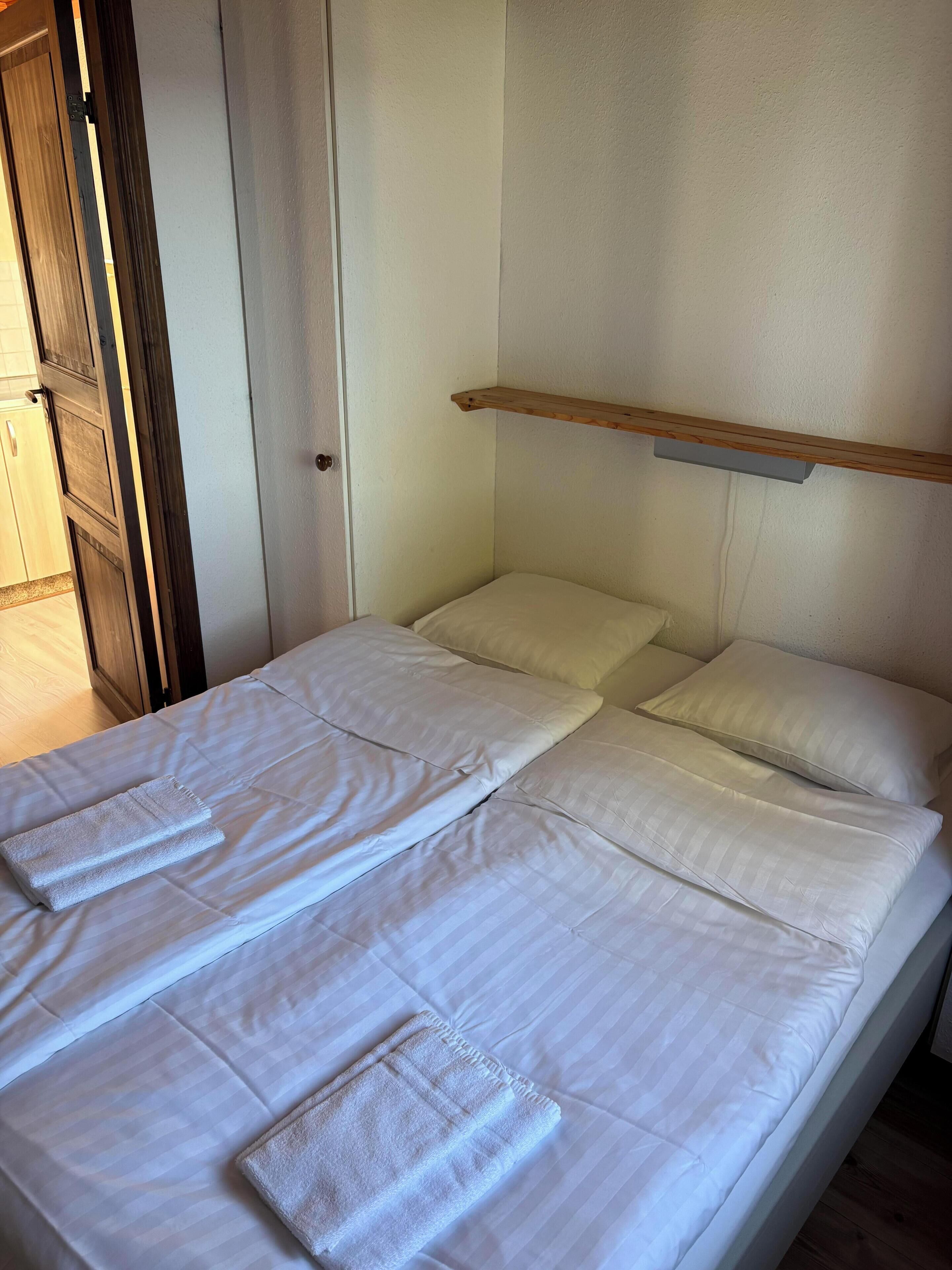 En säng med vita lakan och två kuddar, en dörrkarm i trä och en trähylla ovanför sängen.
