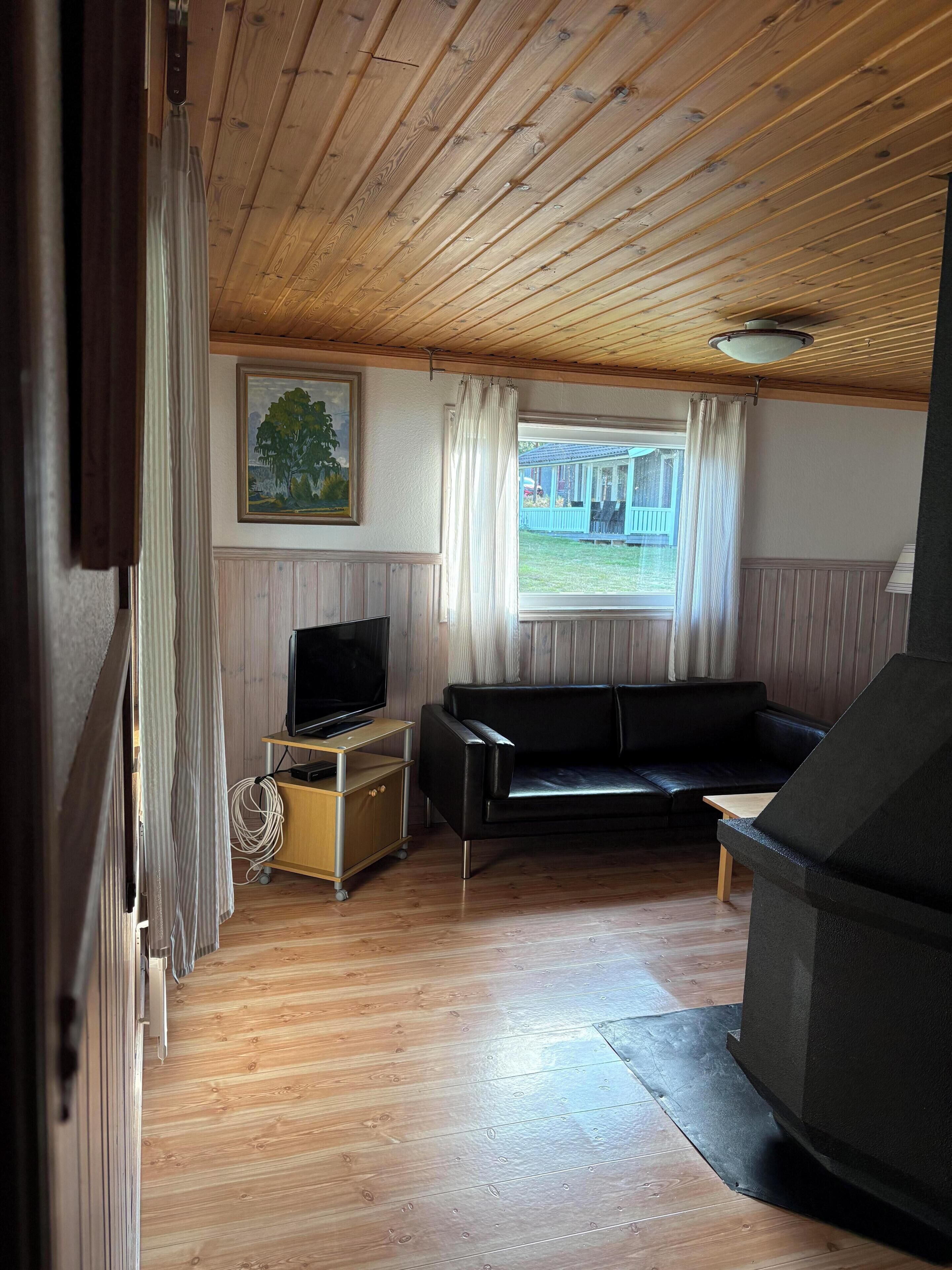 A room with a wooden ceiling, a black leather sofa, a television on a wooden stand, and a window with curtains.