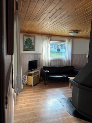 A room with a wooden ceiling, a black leather sofa, a television on a wooden stand, and a window with curtains.