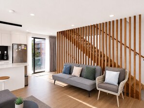 A modern living space with a sofa, a wicker chair, a kitchen area, and a wooden staircase. - Townhouse in Pakuranga (Auckland)