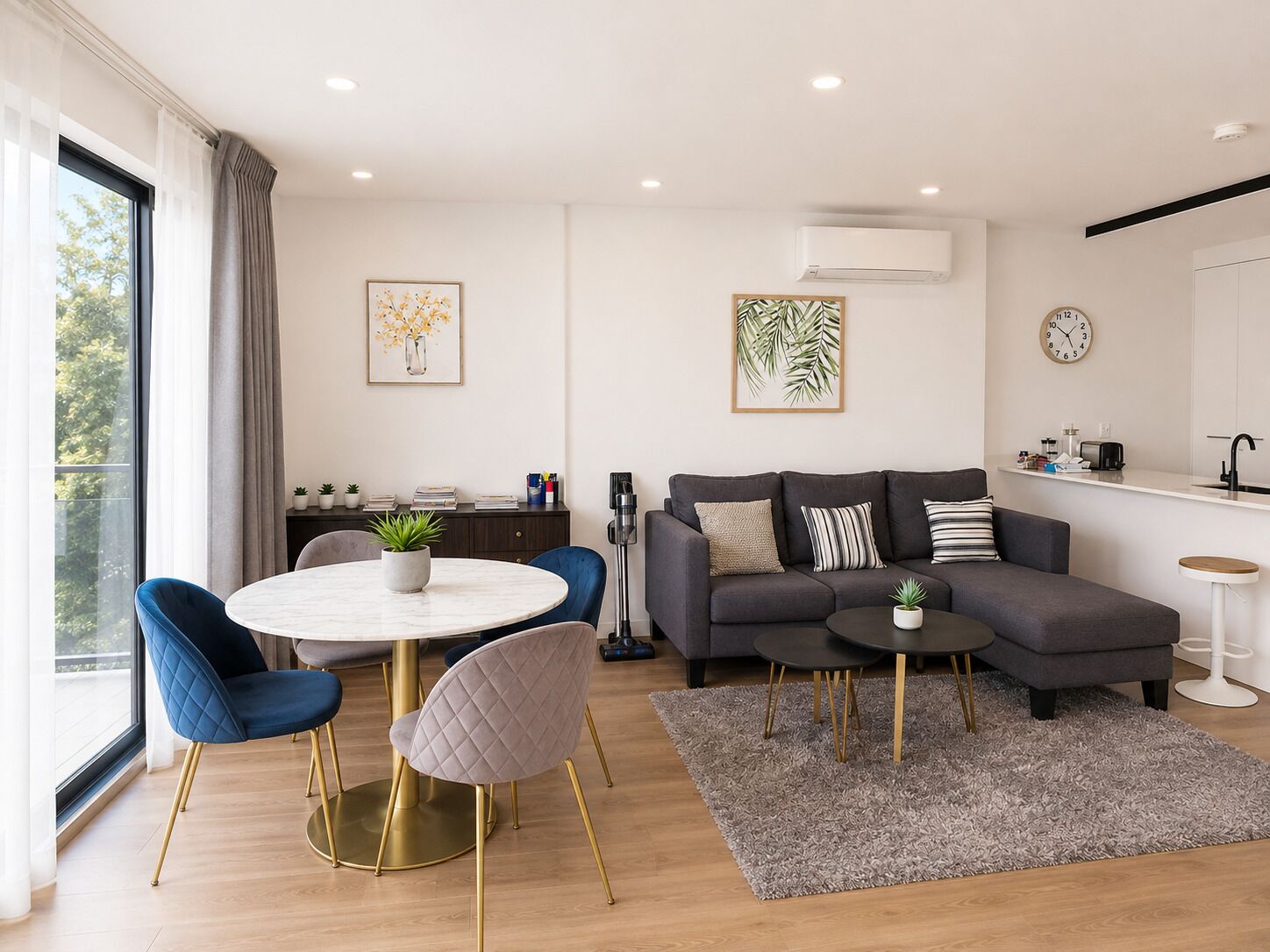 A modern living room with a sofa, a dining table with chairs, a kitchen area, and a large window with curtains.
