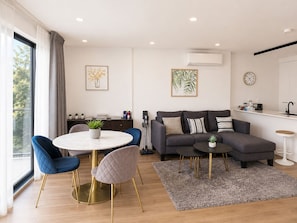 A modern living room with a sofa, a dining table with chairs, a kitchen area, and a large window with curtains.