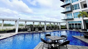 A swimming pool with lounge chairs and tables, surrounded by a building with balconies and windows.