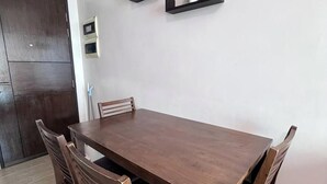 A dining table with four chairs, a dark wooden floor, and a white wall with a small shelf.