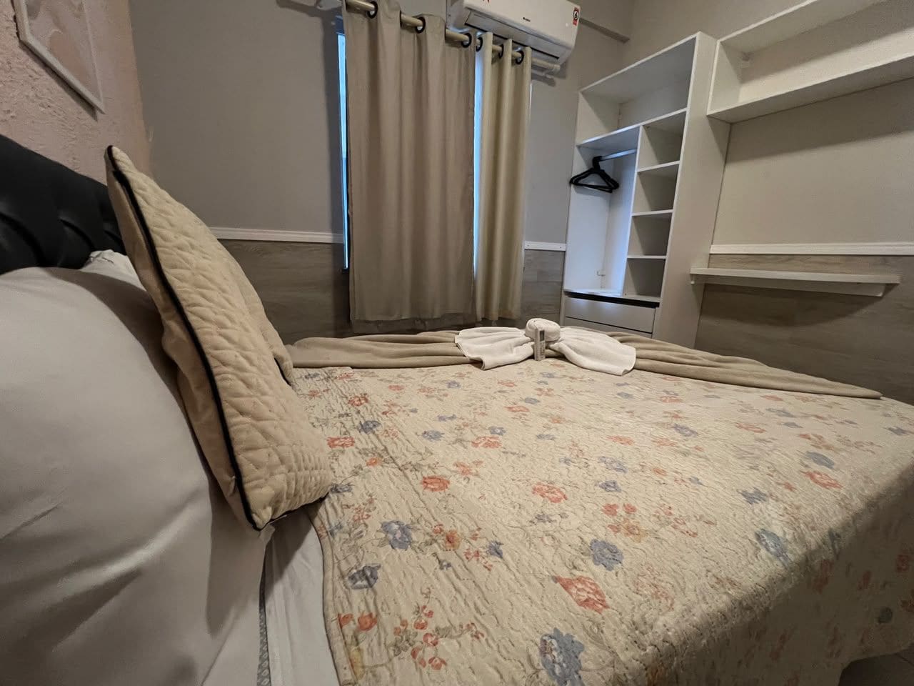 Una cama bien hecha con una colcha estampada, un cojín color beige y una toalla blanca. Se ve una ventana con cortinas color beige, un armario blanco y una estantería blanca.