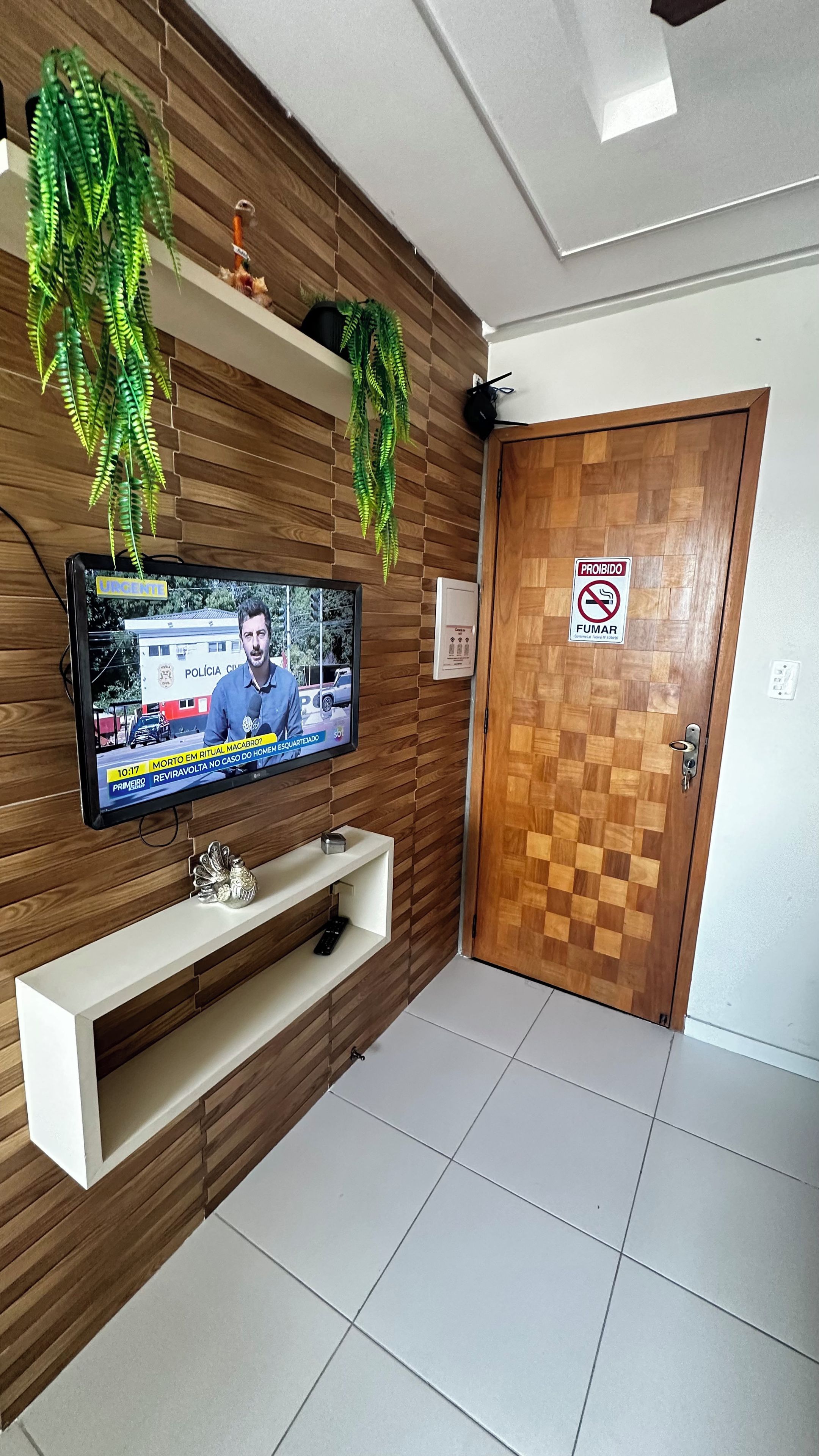A room with a television displaying a news broadcast, a wooden wall with hanging plants, a white console table, and a door with a no-smoking sign.