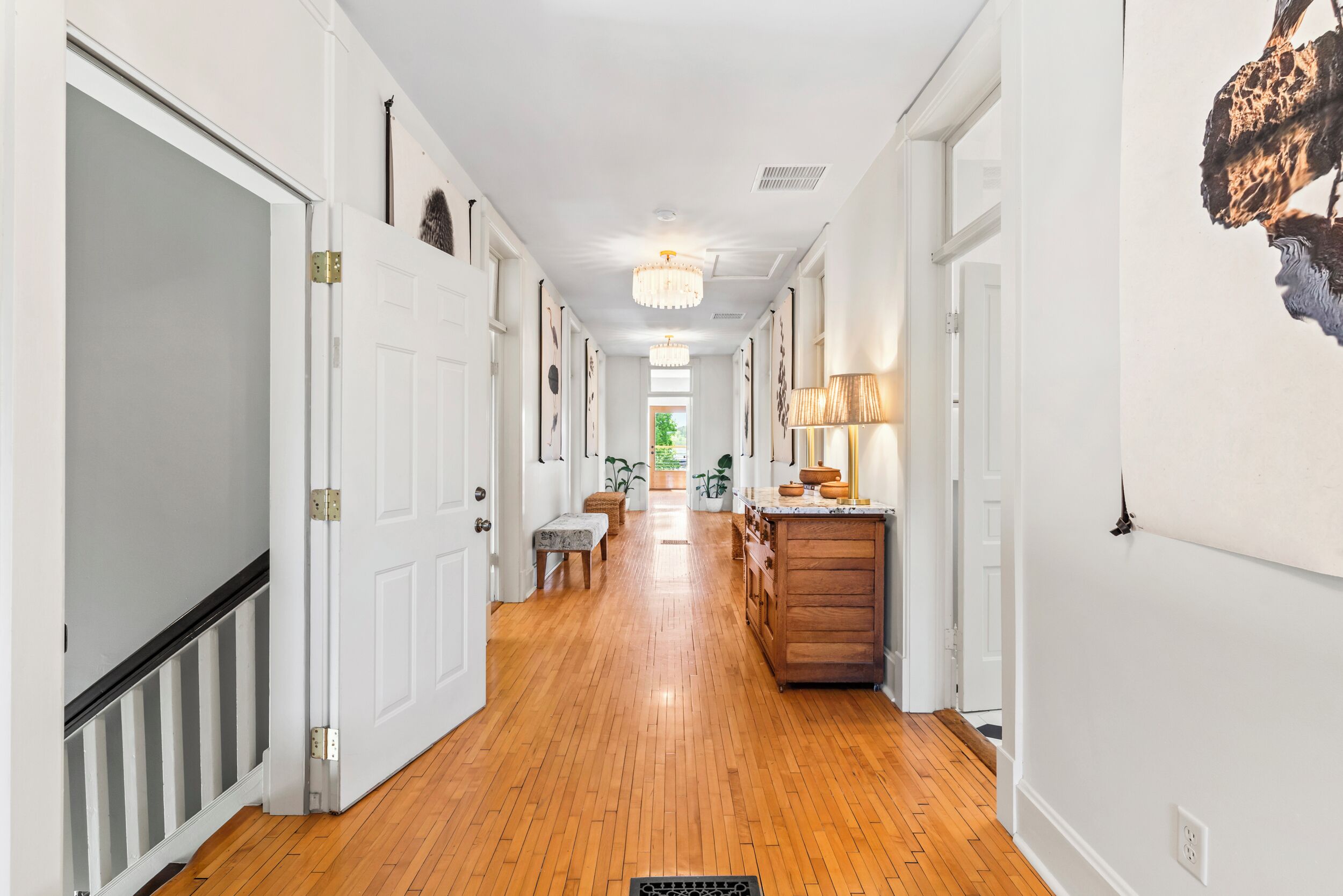 A hallway with wooden flooring, white doors, a bench, and a kitchen area with a wooden cabinet and a lamp.