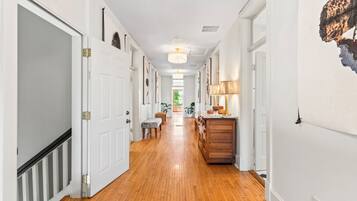 A hallway with wooden flooring, white doors, a bench, and a kitchen area with a wooden cabinet and a lamp.