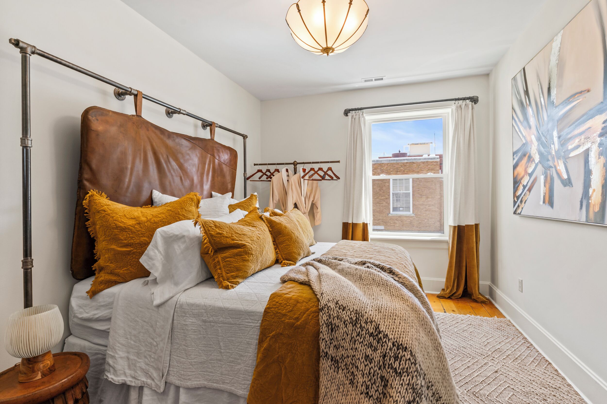 A bedroom with a large bed, a hanging light fixture, a window with curtains, and a painting on the wall.