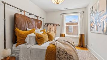 A bedroom with a large bed, a hanging light fixture, a window with curtains, and a painting on the wall.