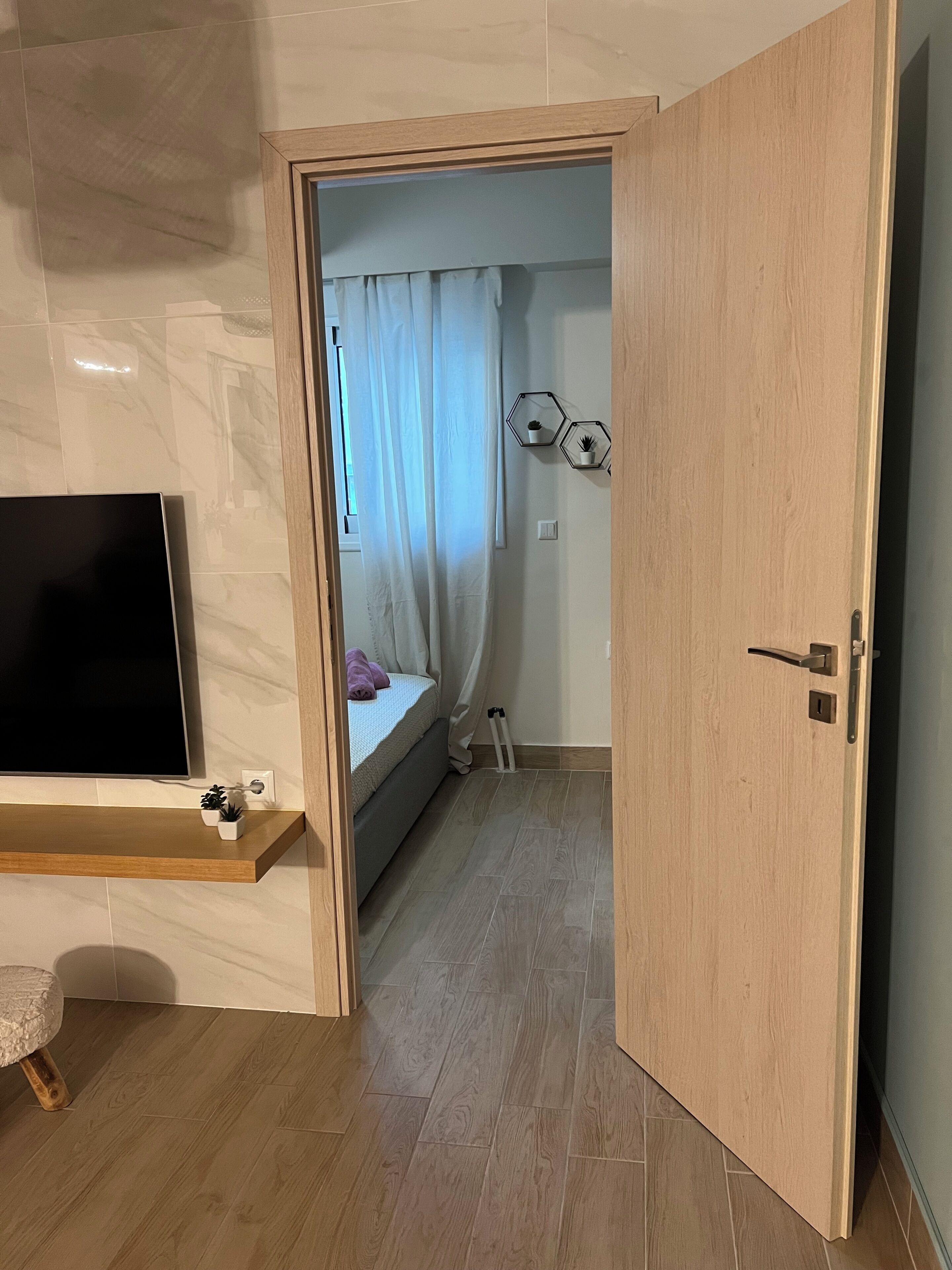 A room with a bed, a TV, a wooden door, and a hallway leading to another room.