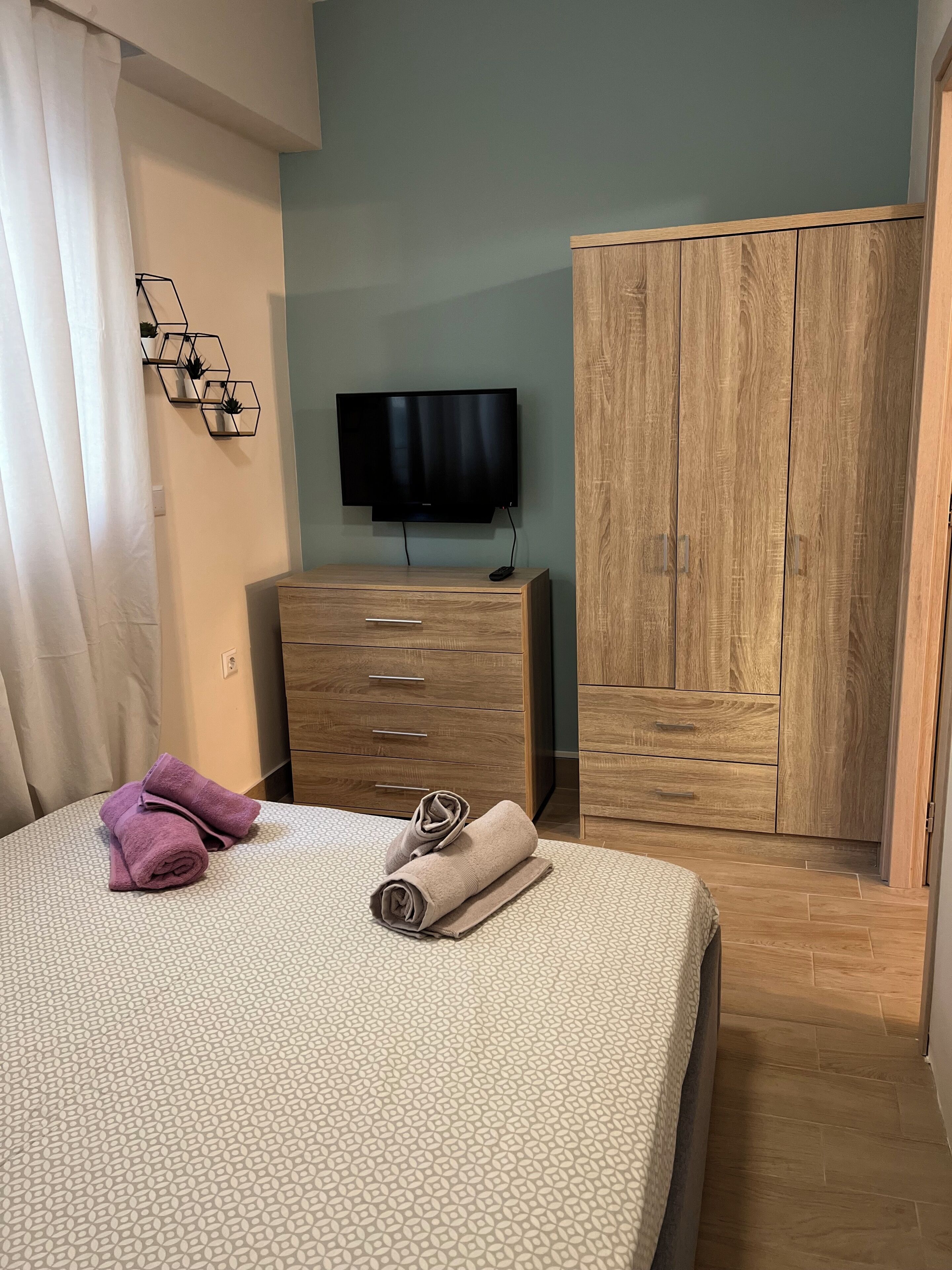 A bedroom with a bed, a TV mounted on the wall, a wooden wardrobe, and a bathroom with a shower curtain.