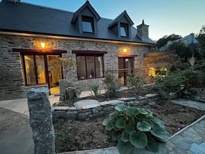 Ein Steinhaus mit geneigtem Dach, großen Fenstern und einem Garten mit Pflanzen und einem Steinweg.