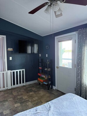 A room with a ceiling fan, a television mounted on the wall, a door with a window, and a shelf with various items.