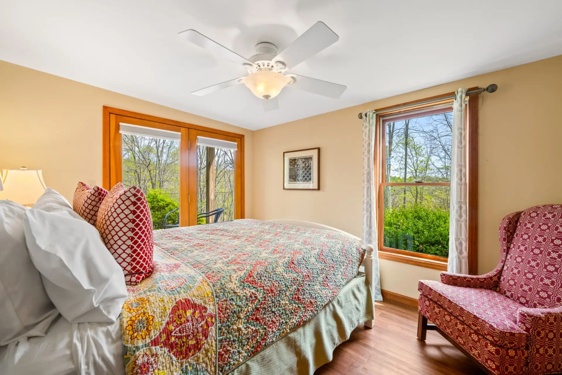 A bedroom with a bed, a chair, a ceiling fan, and a window with a view of trees.