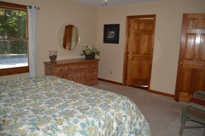 A bedroom with a large bed, a dresser, a mirror, a door, and a window with a view of trees.