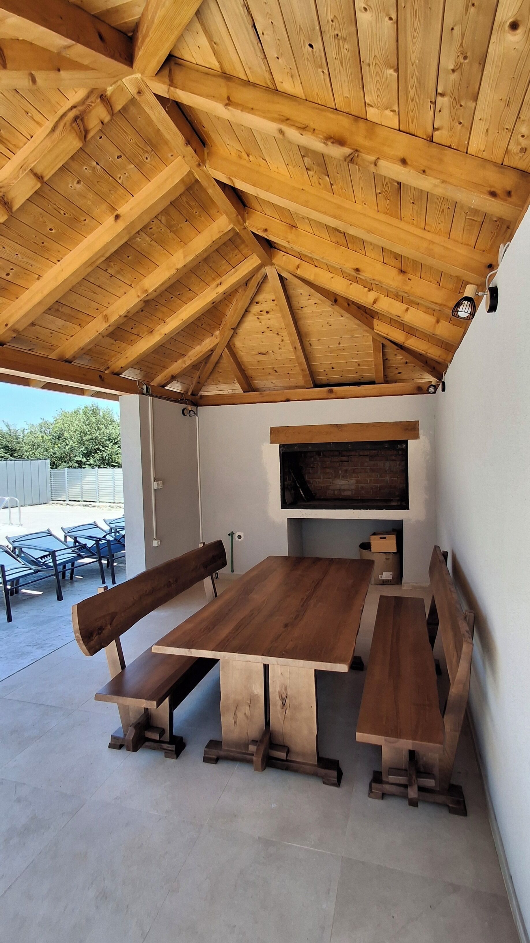 A covered outdoor seating area with a wooden table and benches, a brick fireplace, and a view of the outside.