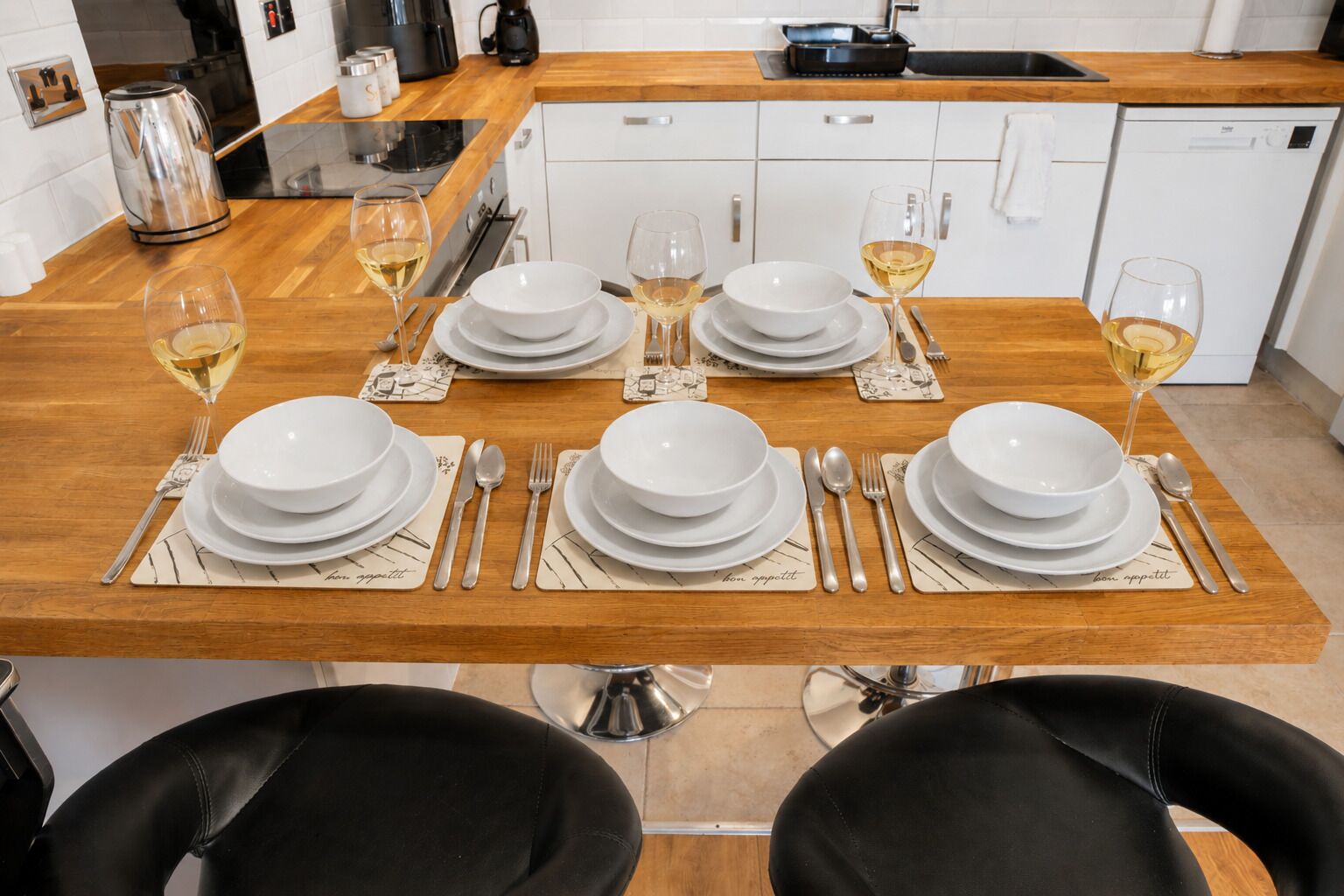 A dining table set with white plates, silverware, and wine glasses, with a coffee maker and kettle on the counter.