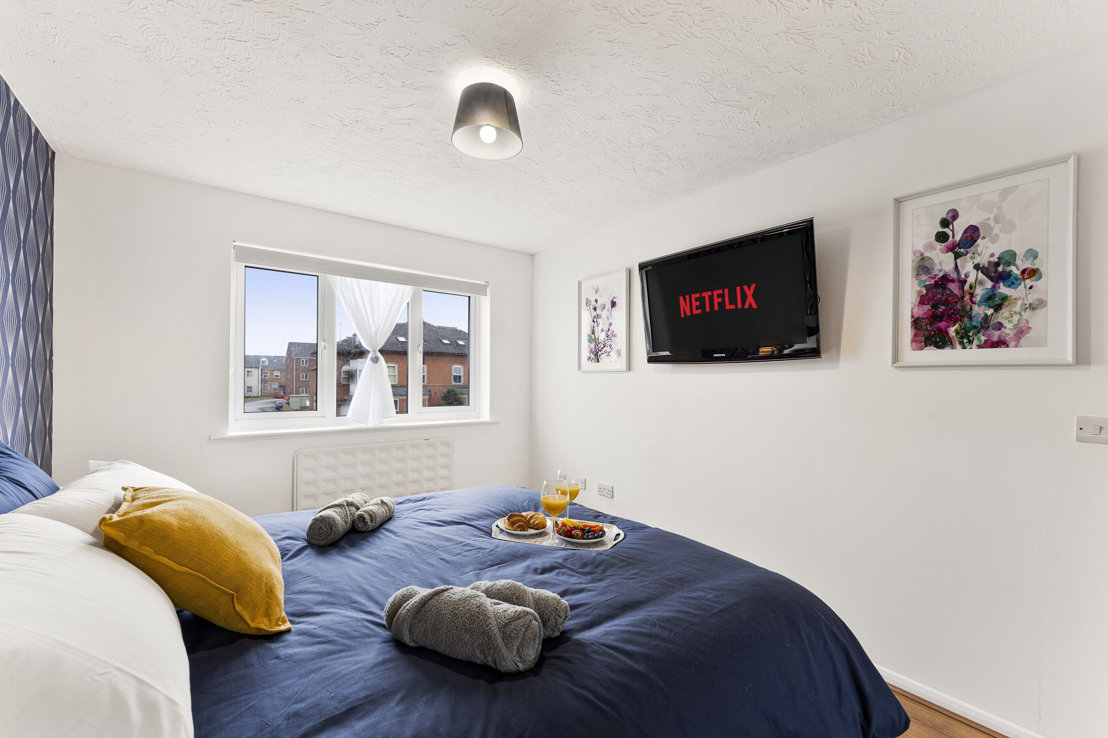 Ein Schlafzimmer mit einem Bett, einem Fenster, einem an der Wand montierten Fernseher und einem gerahmten Bild.