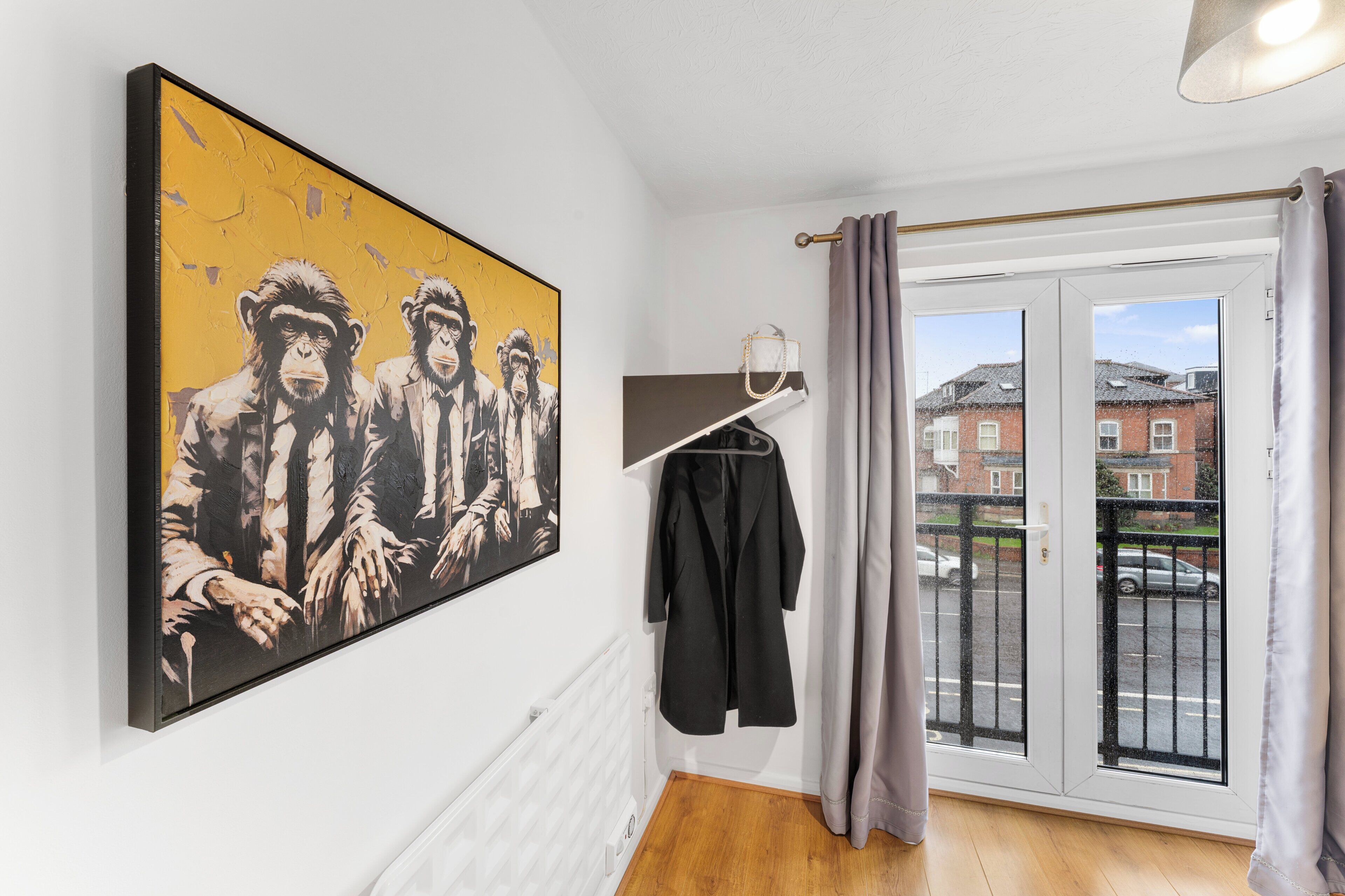 Ein Zimmer mit einem großen Gemälde an der Wand, einem Kleiderständer mit einer Jacke, einem Heizkörper und Blick auf eine Wohngegend durch ein Fenster.