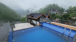 A rooftop swimming pool with a view of a mountainous area and a building labeled 'HOTEL RESTAURANT' in the background.