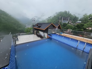 A rooftop swimming pool with a view of a mountainous area and a building labeled 'HOTEL RESTAURANT' in the background.