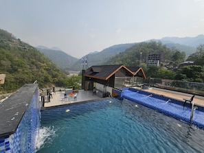 A hotel with a swimming pool, a covered area with seating, and a view of the surrounding hills.