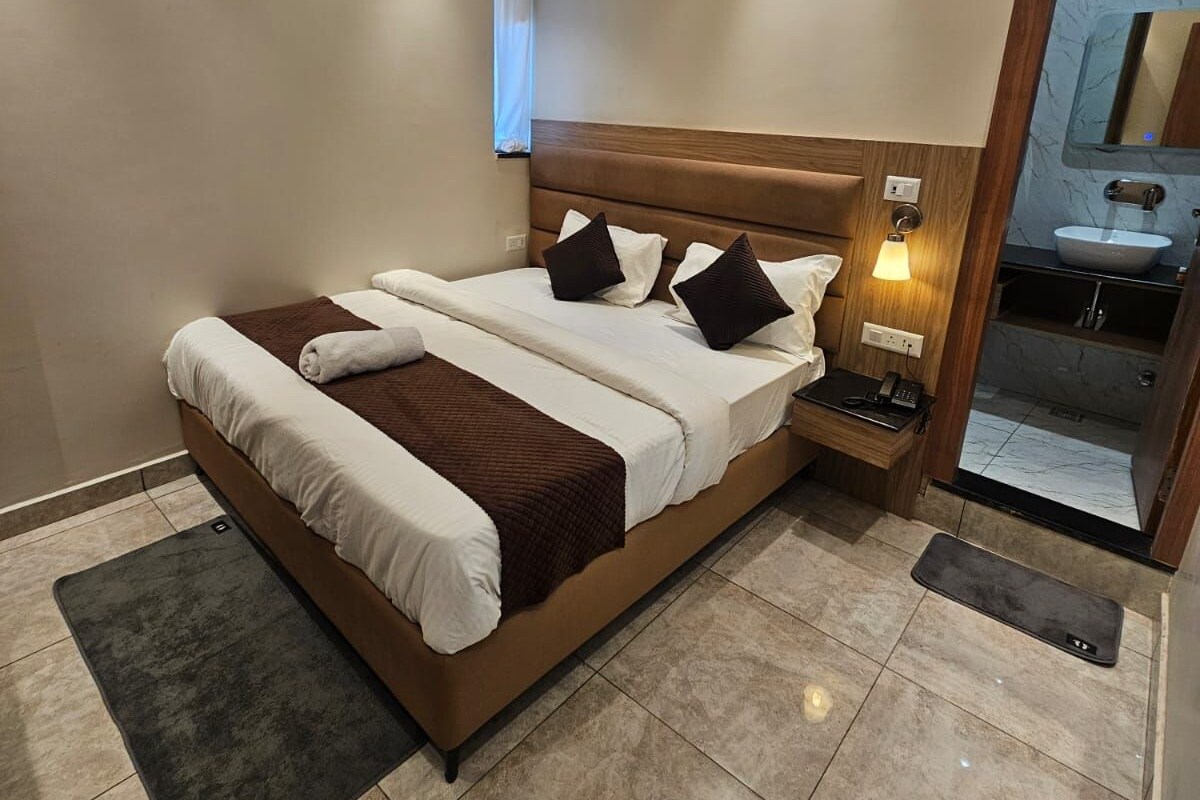 A hotel room with a bed, bedside table, lamp, air conditioning unit, and a bathroom mirror.